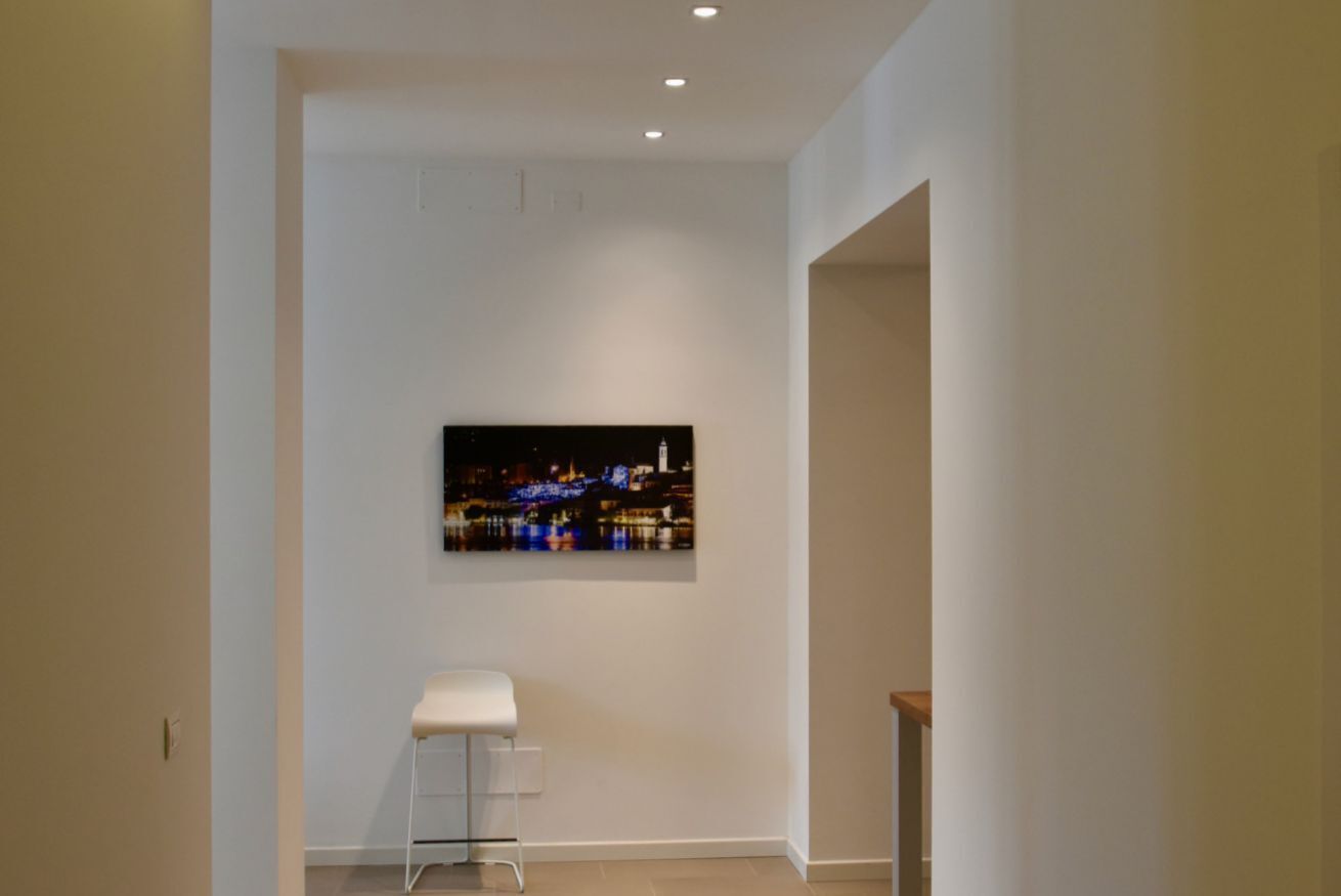 Un corridoio con una parete bianca, un quadro che raffigura un panorama urbano incorniciato e una sedia bianca.