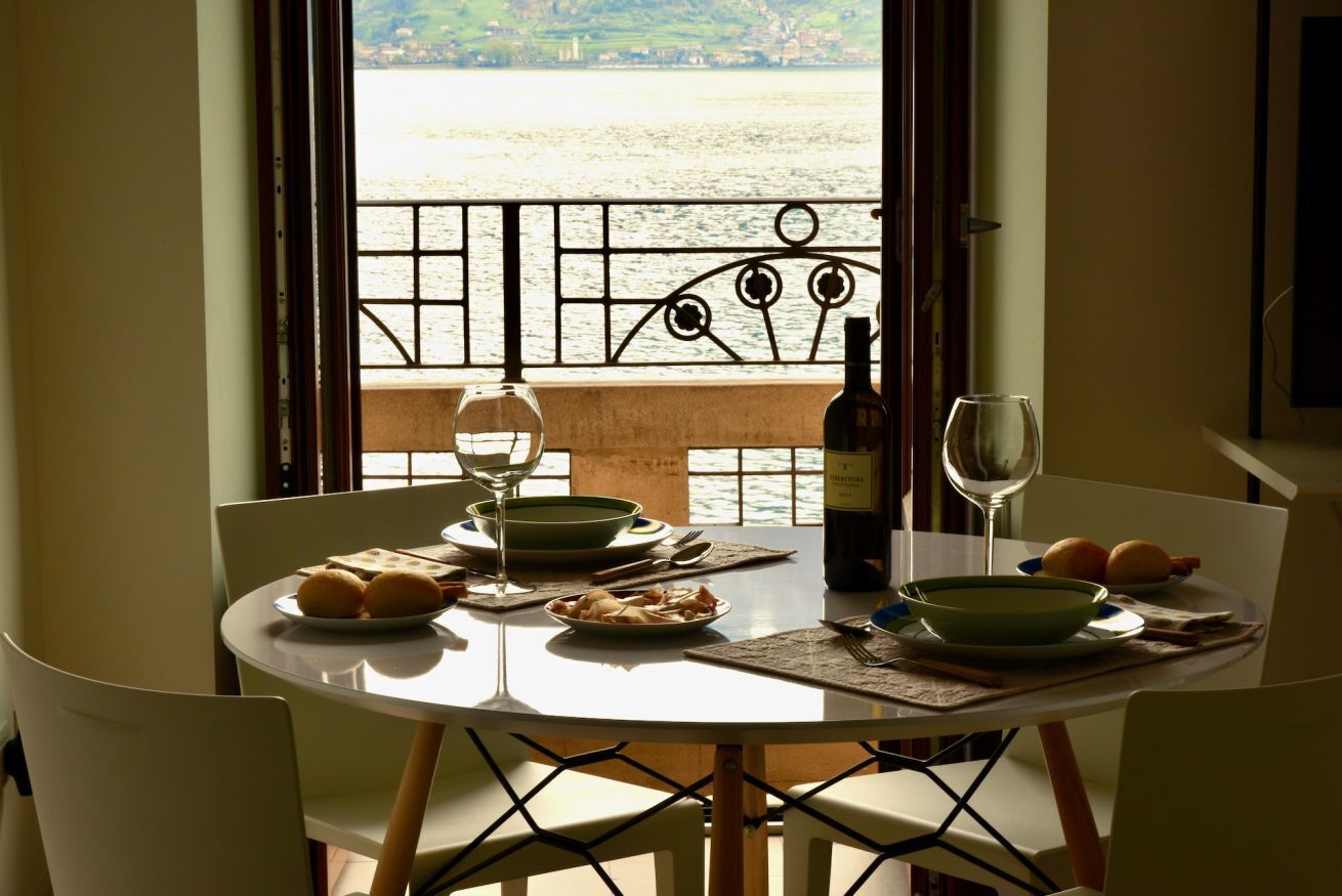 Un tavolo da pranzo con piatti, ciotole e bicchieri da vino, affacciato su una finestra che si apre su un panorama acquatico e un balcone.