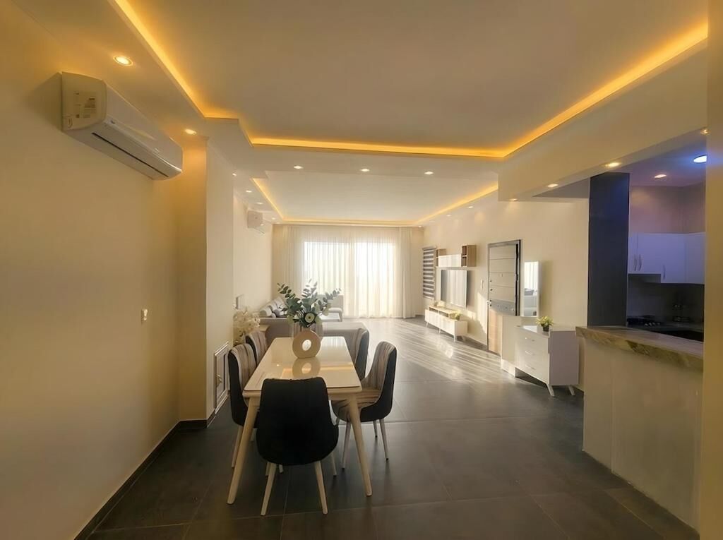 A modern hotel room with a dining area, kitchenette, and a large window with curtains.