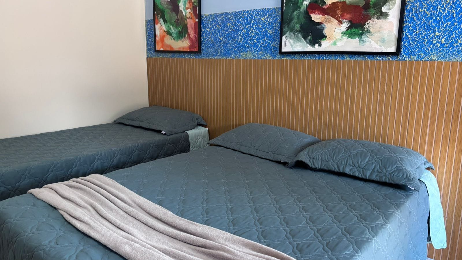 Two single beds with blue bedding, a striped wooden headboard, and abstract art on the wall.