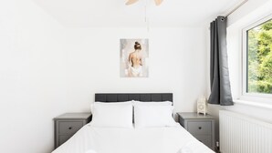Un dormitorio con una cama, dos mesitas de noche, una mesilla auxiliar, una ventana con cortinas y un cuadro colgado en la pared.