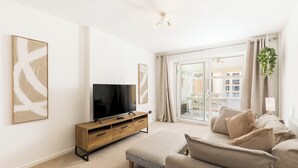Una sala de estar con un televisor de pantalla plana, un mueble bajo de madera, un sofá color beige y un ventanal con cortinas.