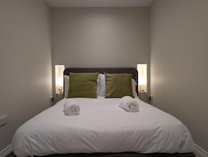 Ein ordentlich bezogenes Bett mit weißen Bettwäsche und zwei grünen Kissen, ein dunkles Kopfteil sowie Nachttischlampen.