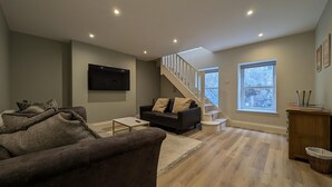 A modern living room with a sofa, a coffee table, a TV mounted on the wall, a staircase, and a window. - Cullercoats Coastal Escape - 3 bedroom Modern Home - Pass the Keys (Newcastle)