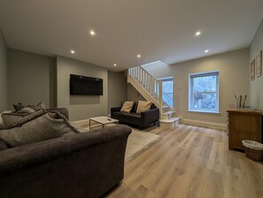 A modern living room with a sofa, a coffee table, a TV mounted on the wall, a staircase, and a window. - Cullercoats Coastal Escape - 3 bedroom Modern Home - Pass the Keys (Newcastle)