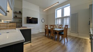 A modern kitchen with a dining area, wooden chairs, a TV, and a clock on the wall. - Cullercoats Coastal Escape - 3 bedroom Modern Home - Pass the Keys (Newcastle)
