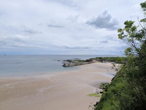 En strand vid kusten med en stenig strandlinje, en liten byggnad nära vattnet och en person som går på sanden.