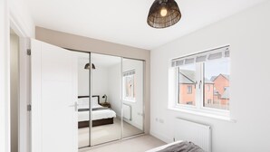 Um quarto com uma cama, mesa de cabeceira, abajur, janela com vista para edifícios e guarda-roupa com espelho.