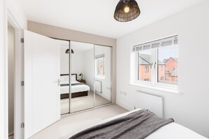 A bedroom with a bed, a bedside table, a lamp, a window with a view of buildings, and a wardrobe with a mirror. - 2 Bed Modern Terrace, Leamington Spa - Pass the Keys (Leamington Spa)
