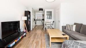 Uma sala de estar moderna com televisão, uma mesa de centro de madeira, um sofá e uma cozinha ao fundo.