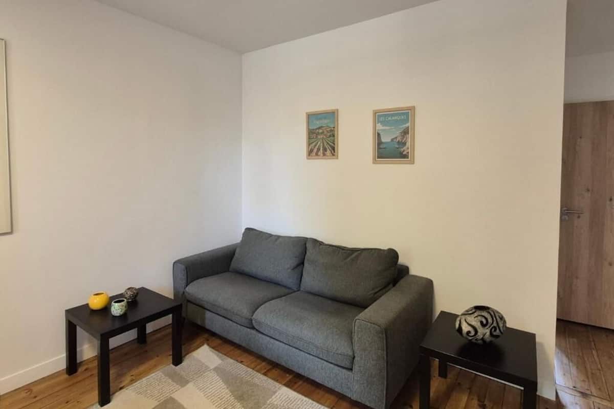 A living room with a grey sofa, two small black tables, a wooden floor, and two framed pictures on the wall.