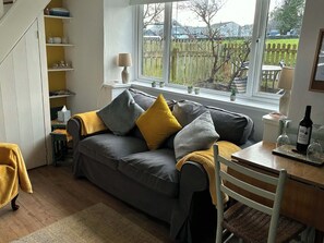 Een gezellige woonkamer met een grijze bank, gele kussens, een houten eettafel en een raam met uitzicht op een omheining en bomen.