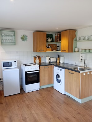 Een compacte keuken met een magnetron, oven en wasmachine, houten kastjes en een betegelde spatwand.
