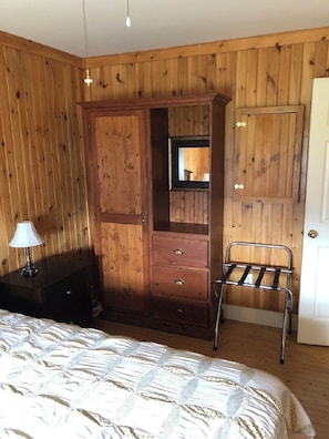 Ein Schlafzimmer mit einem Holzkleiderschrank, einem Spiegel, einem Metallstuhl, einem Nachttisch mit Lampe und einem Bett mit gemustertem Überwurf.