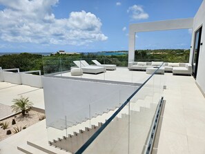A rooftop terrace with a glass railing, white outdoor furniture, and a clear view of the ocean and greenery.