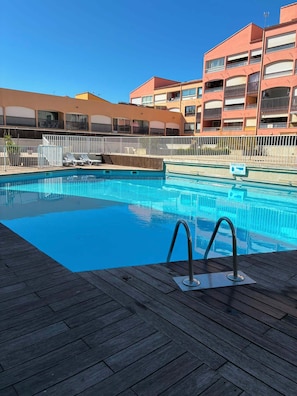 Une piscine dotée d’une terrasse en bois, entourée d’une clôture blanche, et située à côté d’un immeuble à plusieurs étages doté de balcons.