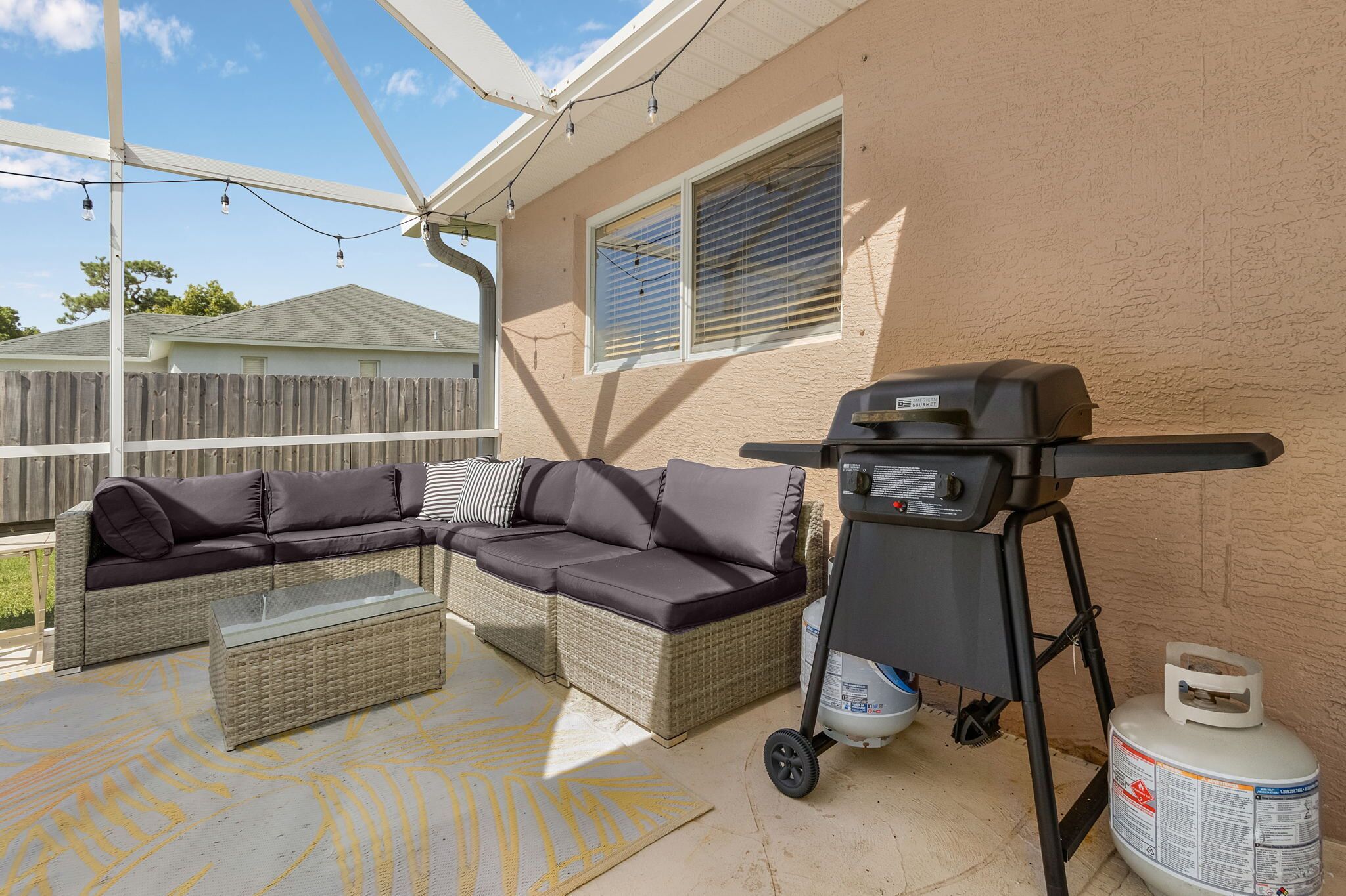 A covered patio with a wicker sofa, a glass coffee table, a grill, and a propane tank.