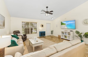 A living room with a sofa, coffee table, TV, ceiling fan, and a window with blinds. - Game Room! Large Pool + Privacy Fence! (Cape Coral)