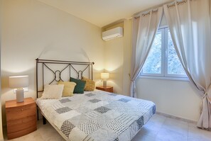 Ein Schlafzimmer mit Bett, Nachttischlampen, einem Nachttisch, einem Fenster mit Vorhängen und einer Klimaanlage.