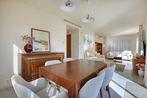 Ein modernes Wohnzimmer mit Esstisch aus Holz, weißen Stühlen, einer Couch und einem Fernseher.