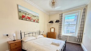 Ein Schlafzimmer mit Bett, Nachttisch, Kommode, Spiegel, einem Fenster mit Vorhängen und einem Bild an der Wand.