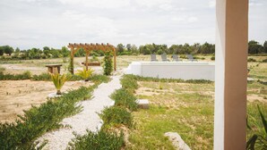 A garden with a pergola, a pathway, and a white wall with lounge chairs.