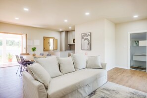 A living room with a sofa, a dining table, chairs, a kitchen area, and a sliding door leading to a bathroom.