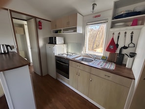 A compact kitchen with a microwave, stove, sink, and wooden cabinets. - 2-Bedroom Mobile Home in the Heart of Nature (Quimper)