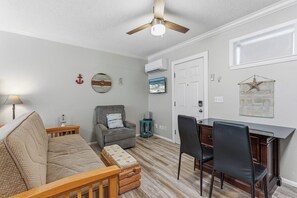A living room with a sofa, a wooden bench, a gray armchair, a dining table with chairs, a ceiling fan, and a TV on the wall.