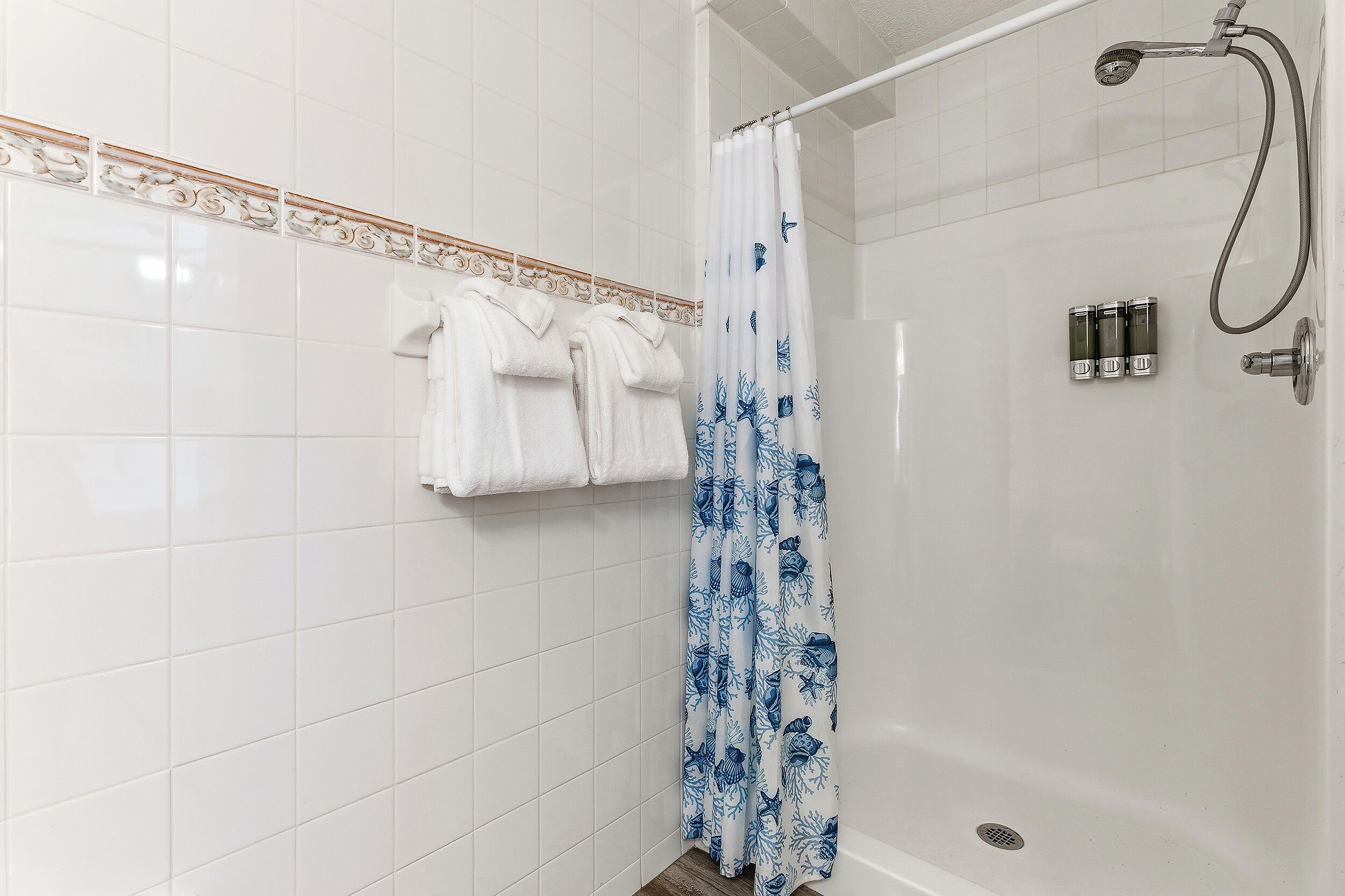 A shower area with a white tiled wall, a shower curtain with a blue floral pattern, and a set of white towels hanging on the wall.