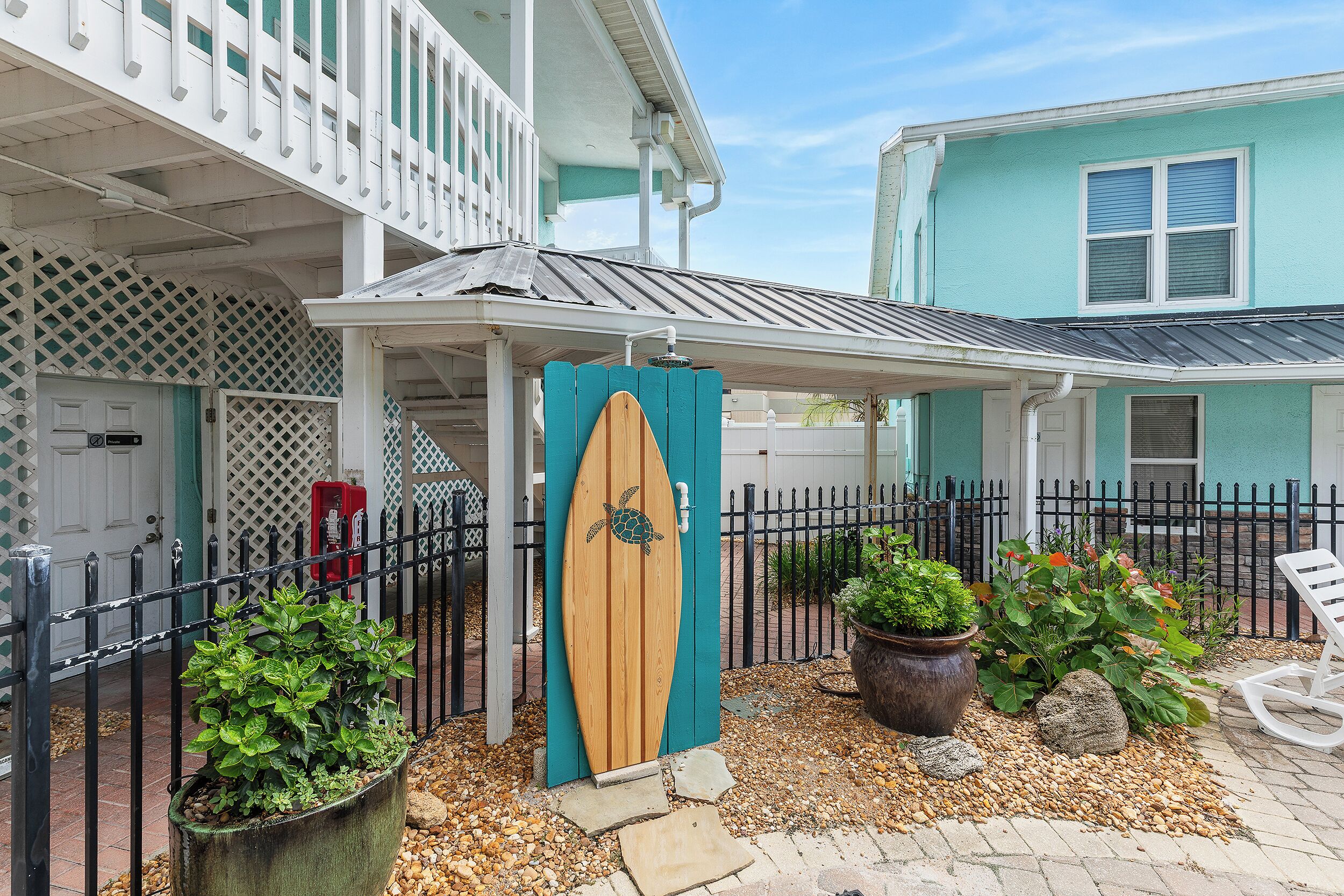 A coastal-style building with a covered porch, a wooden surfboard door, and a fenced outdoor area with potted plants and a chair.