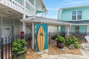 A coastal-style building with a covered porch, a wooden surfboard door, and a fenced outdoor area with potted plants and a chair.