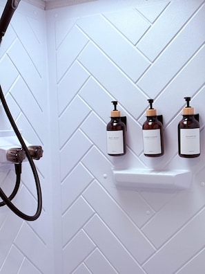Ein Badezimmer mit einer Wand in Weiß mit Fischgrätmuster, drei Flaschen Duschgel, Conditioner und Shampoo sowie einem Duschkopf.