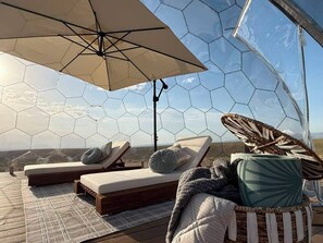 Eine sechseckige Glaskonstruktion mit einer Lounge-Ecke, ausgestattet mit zwei Liegen, einem großen Sonnenschirm und einem gewebten Korb.