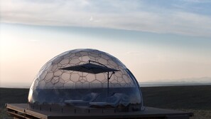 Eine geodätische Kuppel mit einer transparenten Dachkonstruktion, einem Lounge-Bereich mit Sesseln und einem Holzdeck.