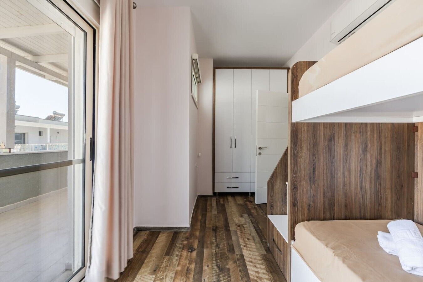 Una habitació amb sòl de fusta, un llit superposat, un armari blanc i una porta corredissa de vidre.