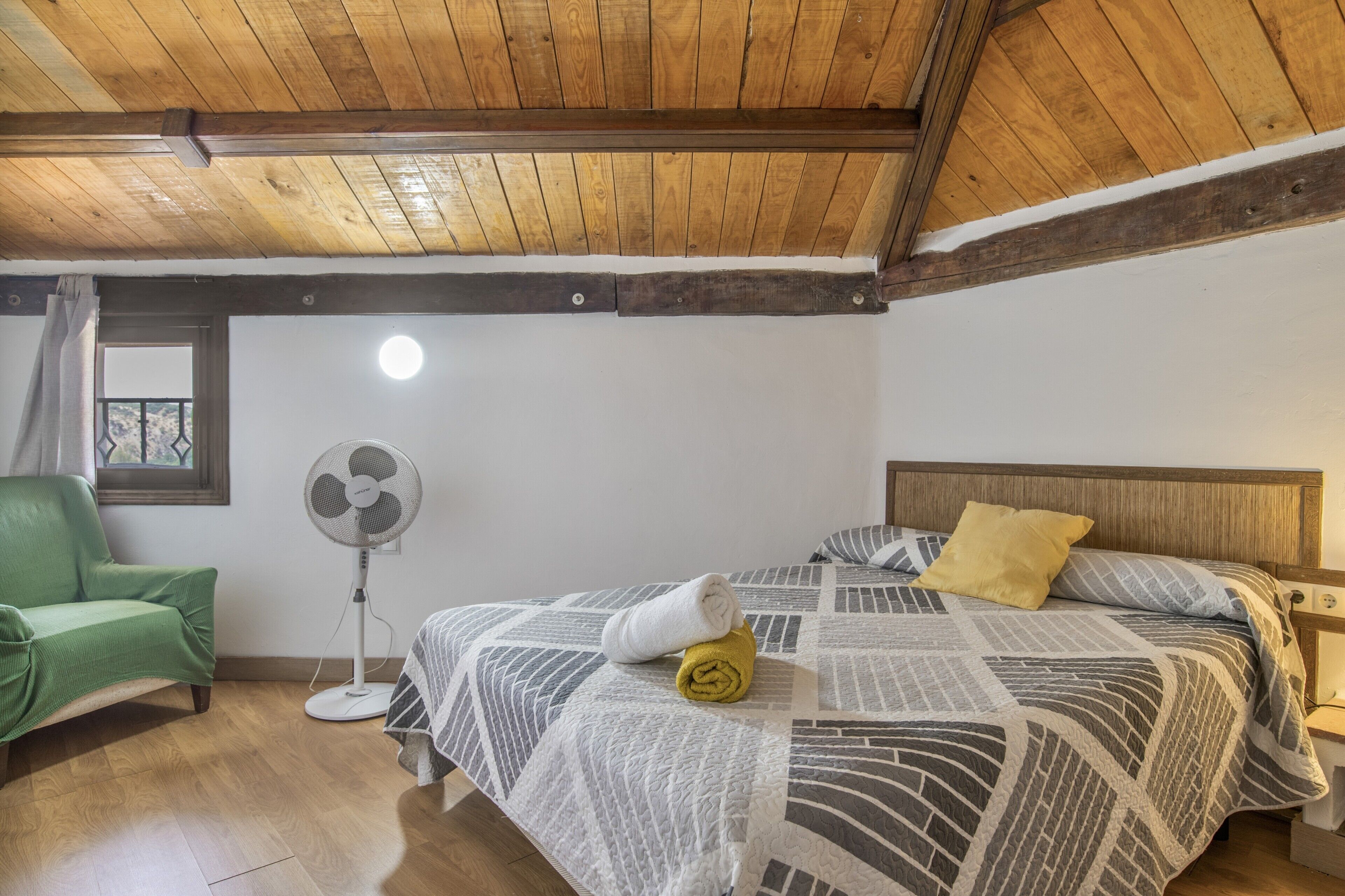 A room with a wooden ceiling, a bed with a patterned bedspread, a green chair, a standing fan, and a window with curtains.