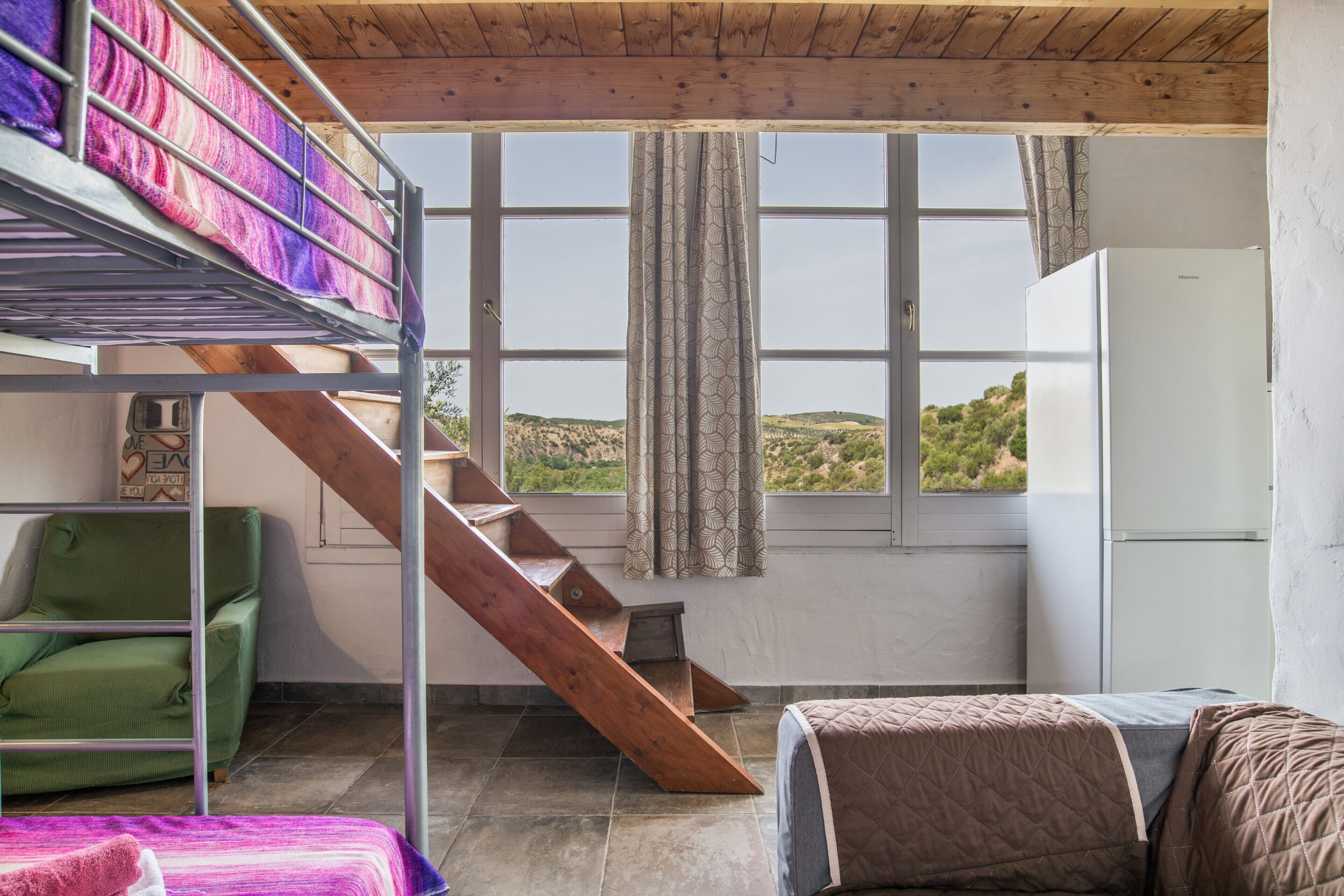 Una habitación con literas, una escalera, una silla verde, una cama con colcha y vistas a las colinas desde una ventana.