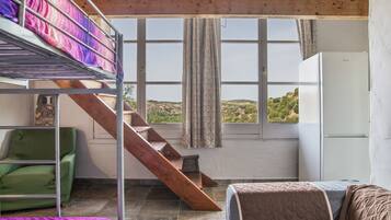 Una habitación con literas, una escalera, una silla verde, una cama con colcha y vistas a las colinas desde una ventana.