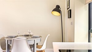 Een eethoek met een ronde tafel, witte stoelen, een zwarte vloerlamp en een raam met jaloezieën.