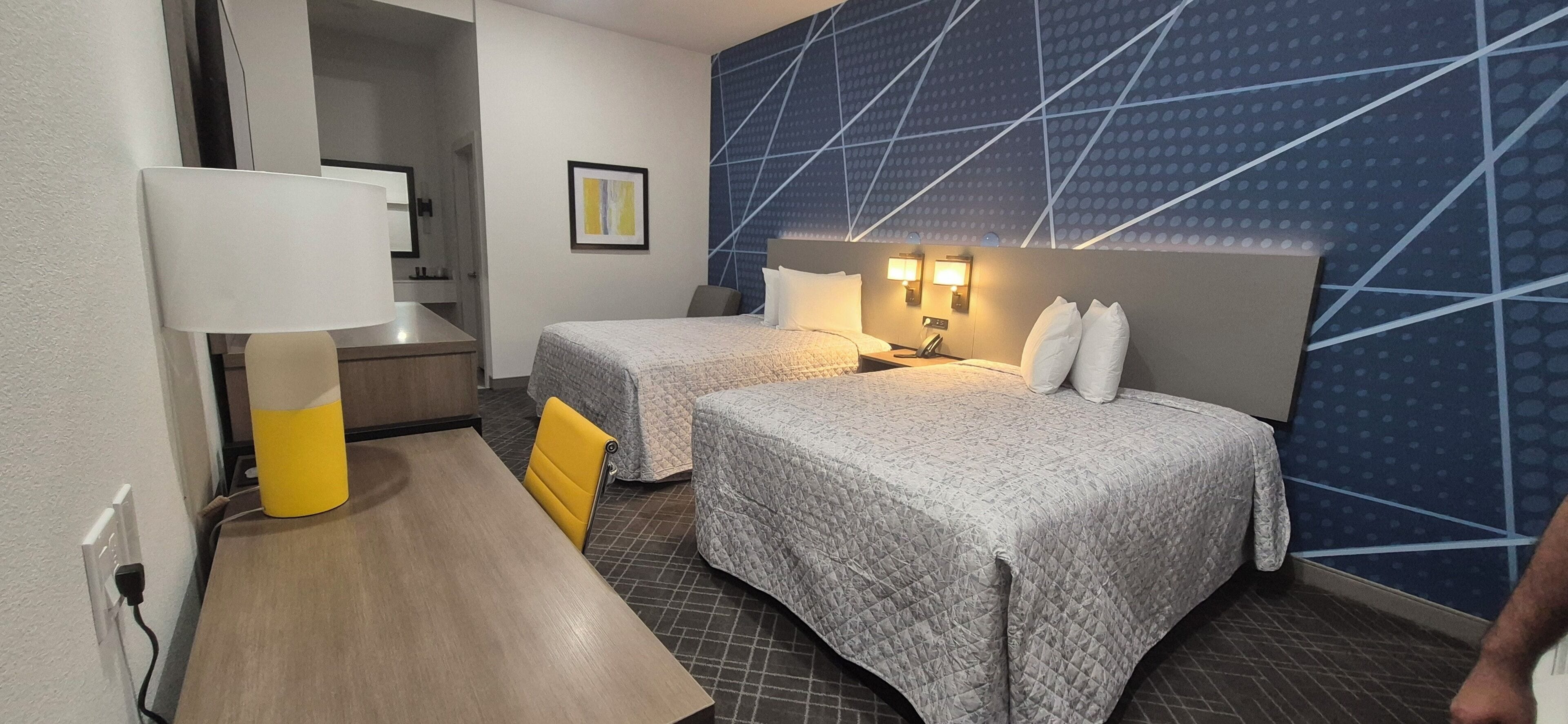 A hotel room with two beds, a desk, a lamp, and a wall with a geometric pattern.
