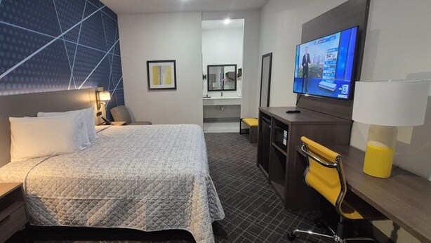 A hotel room with a bed, bedside tables, a TV, a desk, a chair, a mirror, and a bathroom in the background. - Platinum Inn Copperfield (Houston)