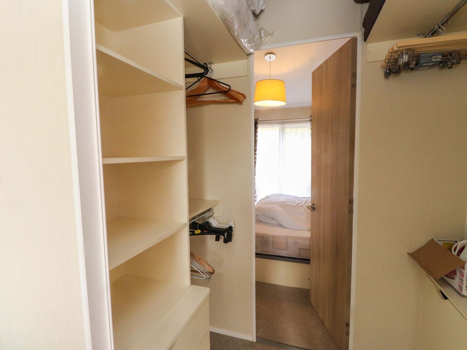 A narrow hallway leading to a bedroom with a bed, a hanging light fixture, and a closet with shelves.