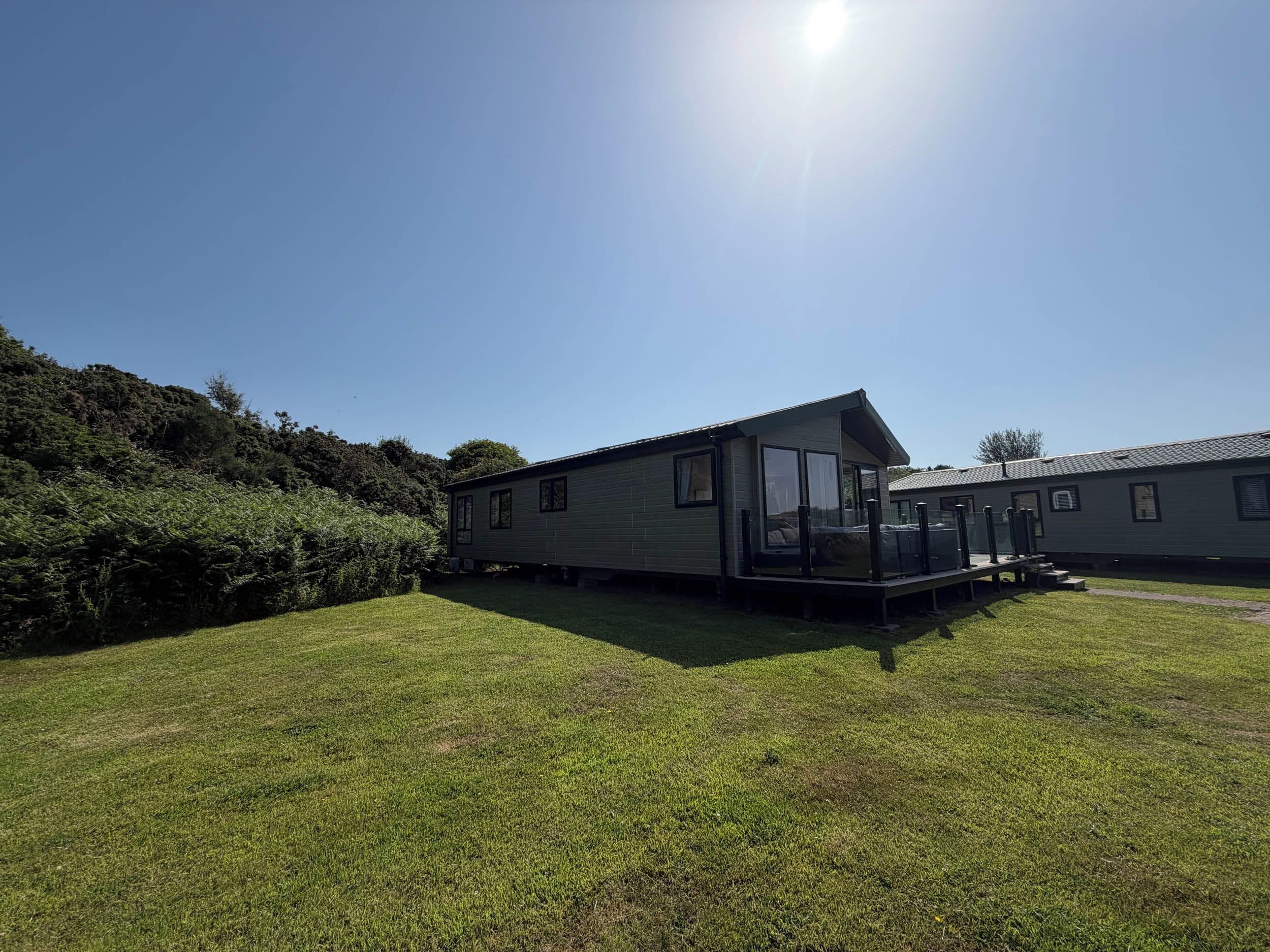A single-story mobile home with a deck, surrounded by a grassy area and dense foliage.