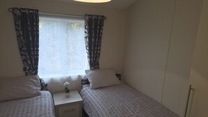 A bedroom with two single beds, a bedside table with a lamp, and a window with curtains. - The Stables (Pwllheli)