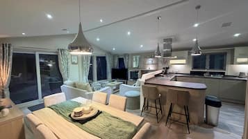 A modern kitchen with a central island, dining area with a table set for two, and a living room with a sofa and TV.