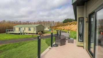 A modern building with a balcony, a pathway, and a grassy area with mobile homes in the background.