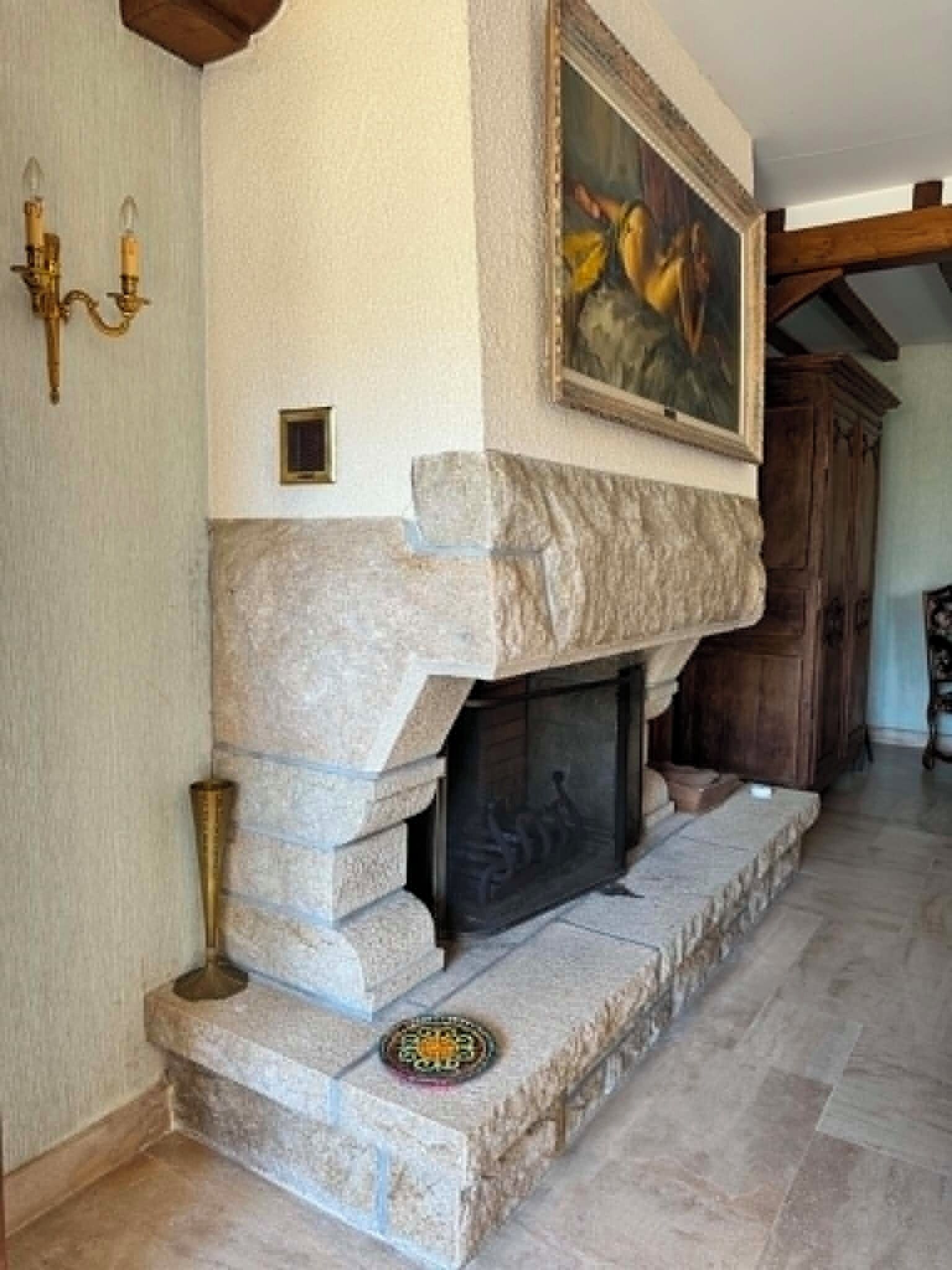 Une cheminée en pierre surmontée d’un tableau, une petite assiette décorative posée sur le foyer, et un meuble en bois à l’arrière-plan.