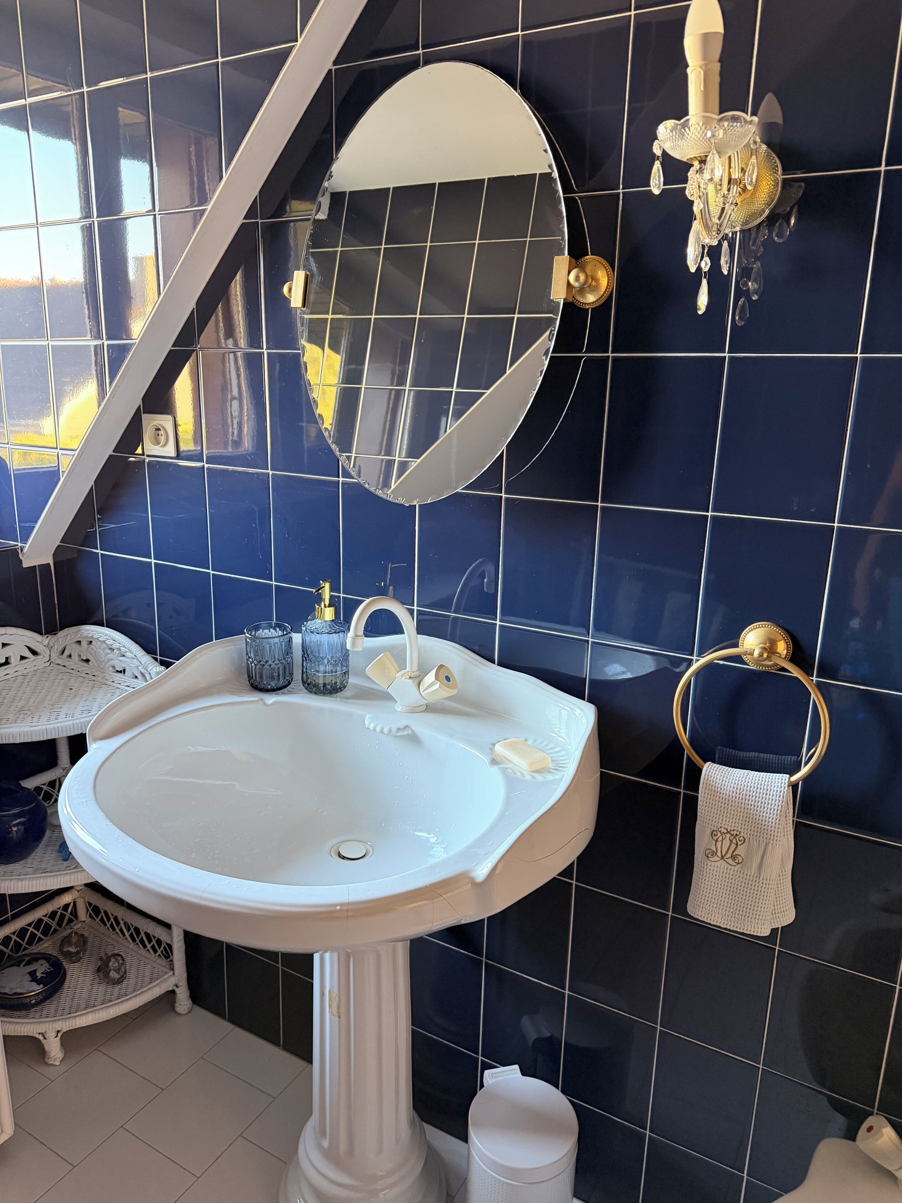 Un baño con lavamanos blanco, espejo, argolla dorada para toallas y una lámpara de pared.