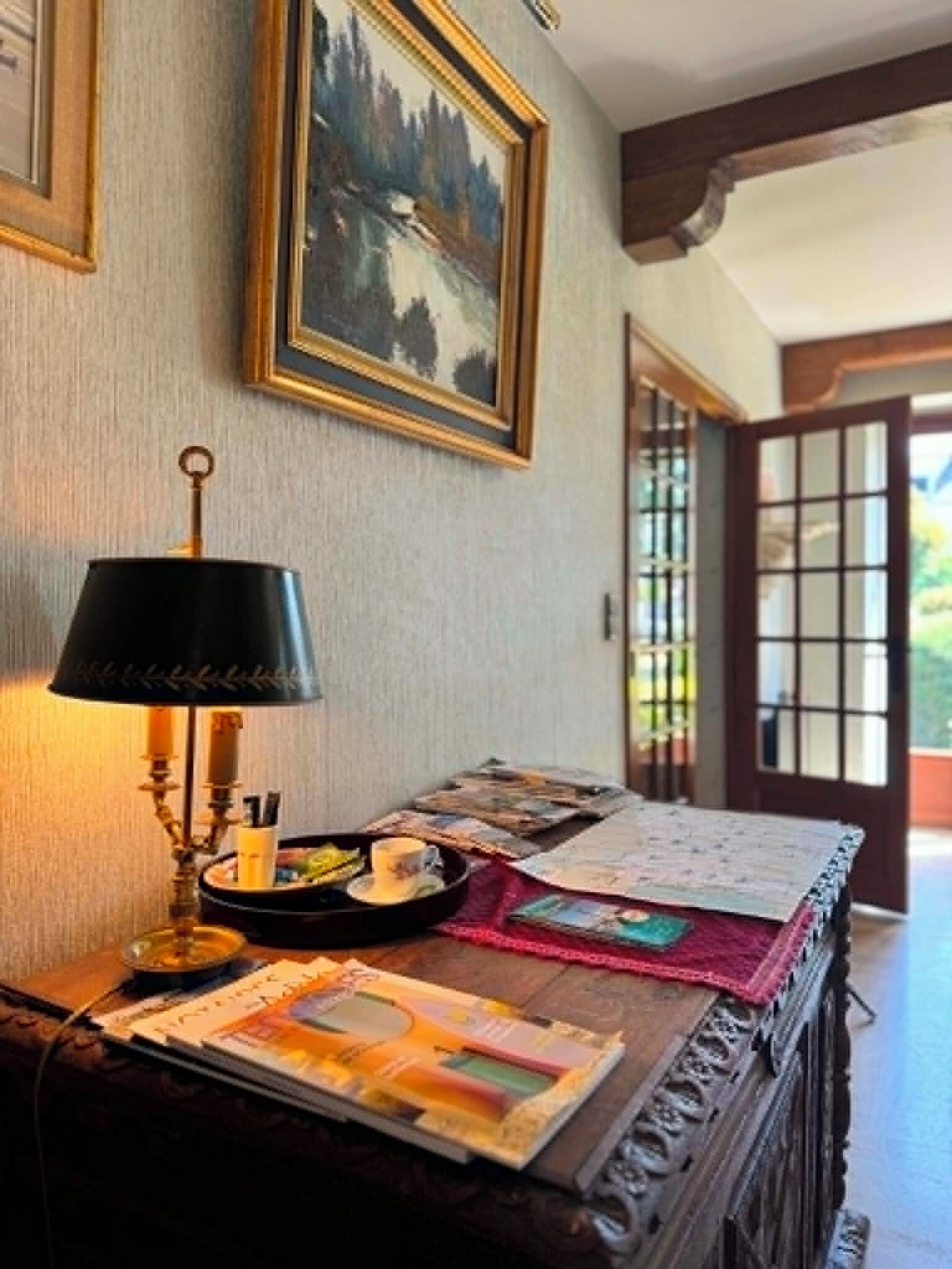 Une pièce avec un bureau, une lampe, un tableau encadré et une vue sur l’extérieur, visible à travers une porte.